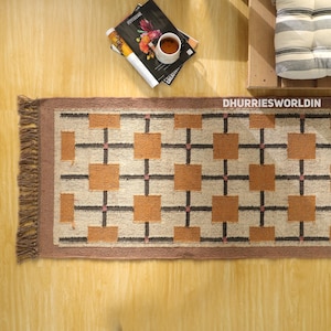 May include: A rectangular woven runner rug featuring a geometric grid pattern of tan squares separated by dark brown lines on a cream background. The rug has a solid brown border and fringed edges on the left side. The text DHURRIESWORLDIN is visible.