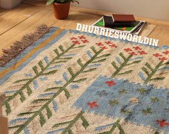 May include: A rectangular woven rug featuring a beige base with repeating green and blue floral patterns and red accents. The rug has a fringed edge and the text DHURRIESWORLDIN is printed in white across the upper portion of the fabric surface.