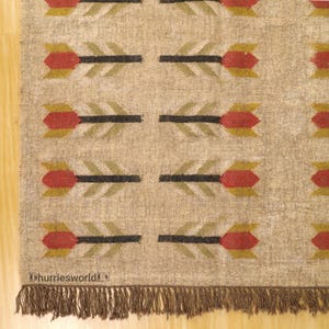 May include: Beige rug with a repeating arrow pattern in red, black, and olive green. The rug has a fringed edge and the text "dhurriesworld" is visible in the bottom left corner.