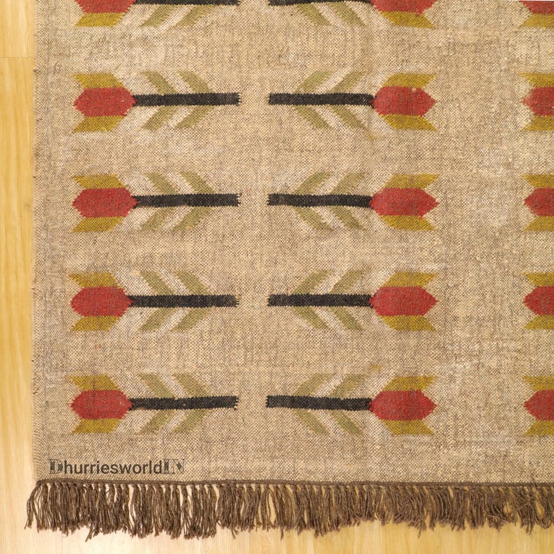 May include: Beige rug with a repeating arrow pattern in red, black, and olive green. The rug has a fringed edge and the text "dhurriesworld" is visible in the bottom left corner.