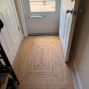 May include: A rectangular area rug crafted from natural tan jute fibers, featuring a braided texture and a concentric border pattern. The rug is positioned on a light-colored tiled floor in an entryway, directly in front of an open white door.