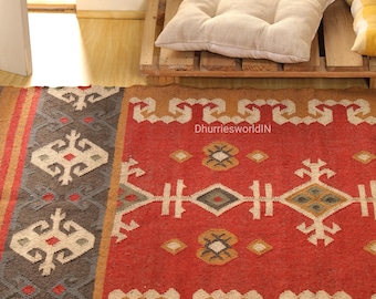 May include: A vibrant red and brown area rug with geometric patterns. The rug features a central design with diamonds, crosses, and floral motifs in white, gray, and yellow. The rug is placed on a wooden floor, with a neutral-colored cushion on a wooden pallet.