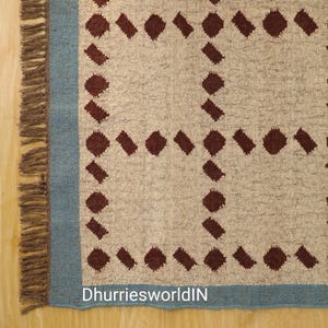 May include: A woven rug featuring a cream-colored base with a grid pattern of dark brown geometric shapes. The rug has a solid light blue border and brown fringe along the left edge. The text DhurriesworldIN is printed in white at the bottom.