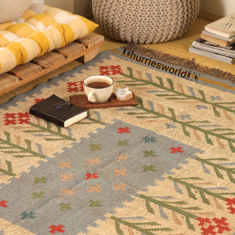 May include: A beige and blue patterned rug with a floral design. The rug has a central blue section with red, green, and orange floral accents. A cup of tea, a small candle, and a book are on the rug.
