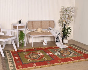 May include: A green, red, and beige handwoven rug with a geometric pattern. The rug is in a living room setting with a white bench and a side table.