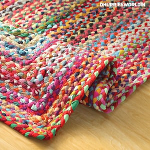 May include: A multi-coloured braided rug featuring a vibrant mix of red, blue, yellow, green, and pink fabric strips woven into a dense, textured pattern. The corner of the rug is folded over, revealing the thick, braided construction. Text reads DHURRIESWORLDIN.