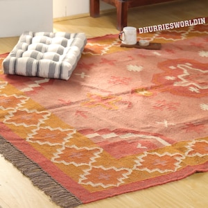 May include: A rectangular area rug featuring a salmon pink base with orange and cream geometric patterns, including diamond shapes and a fringed edge. A white and grey striped tufted floor cushion sits on the rug, with the text DHURRIESWORLDIN visible.