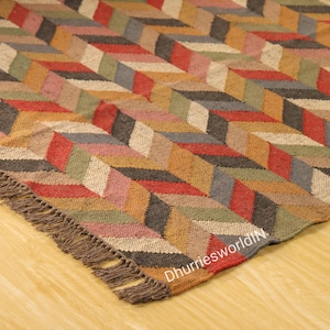 May include: A multi-coloured area rug featuring a geometric design in red, brown, grey, green, and beige. The rug has a fringed edge and a textured appearance, suitable for home decor.