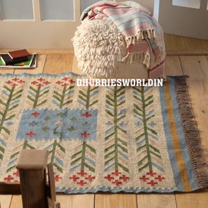 May include: A rectangular woven rug featuring a repeating pattern of green, blue, and beige stylized floral stalks on a cream background, with a blue central rectangular panel, a yellow border, and brown fringe. The text DHURRIESWORLDIN is visible.