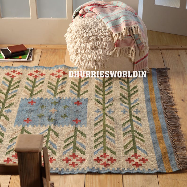 May include: A rectangular woven rug featuring a repeating pattern of green, blue, and beige stylized floral stalks on a cream background, with a blue central rectangular panel, a yellow border, and brown fringe. The text DHURRIESWORLDIN is visible.