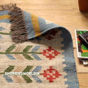 May include: A woven rug featuring a cream-colored base with a repeating pattern of green, blue, and red geometric floral motifs. The rug has a blue border with a yellow stripe and brown fringe along the edge. The text DHURRIESWORLDIN is visible.