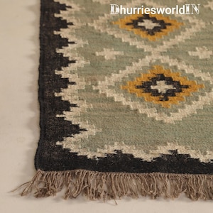 May include: A woven rug featuring a sage green background with a repeating diamond pattern in mustard yellow, black, and off-white. The rug has a thick black border and a fringed edge. The text DhurriesworldIN is visible in the top right corner.