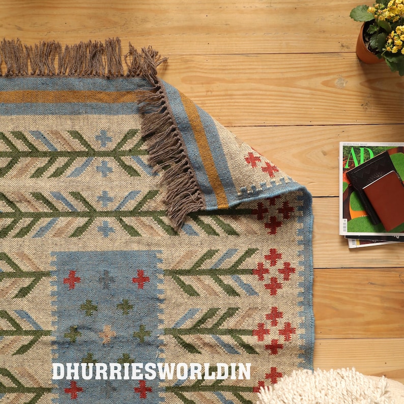 May include: A woven rug featuring a natural tan base with a geometric pattern in blue, green, red, and gold. The rug has a textured surface, a folded corner, and brown fringe along the edges. The text DHURRIESWORLDIN is printed on the rug surface.