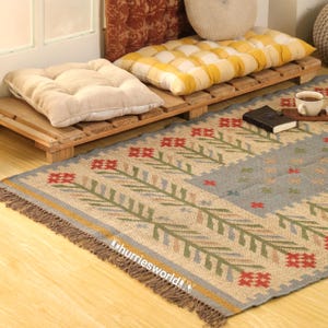 May include: An interior scene with a beige rug with a floral pattern and fringed edges. Two cushions rest on a wooden pallet, one beige and one yellow and white checkered. A small book and a cup of tea are also visible in the scene.