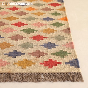 Peut inclure: Un tapis tissé avec une base beige clair présentant un motif répétitif de petites croix géométriques multicolores en rouge, bleu, vert, jaune et rose. Le tapis comprend un bord à franges en bas et le texte DHURRIESWORLDIN.