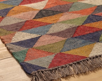 May include: A colorful geometric rug with a diamond pattern in shades of red, orange, blue, green, and brown. The rug has a fringed edge and the text "DHURRIESWORLDIN" is visible in the upper right corner.