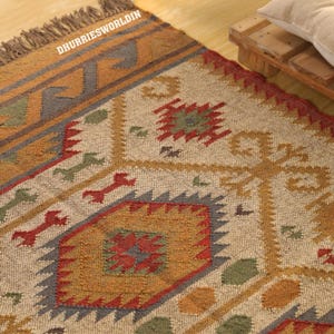 Op de afbeelding: Een geweven vloerkleed met een crèmekleurige basis en geometrische patronen in rood, mosterdgeel, saliegroen en leiblauw. Het kleed heeft een getextureerd oppervlak, een rand met franjes en de tekst DHURRIESWORLDIN in het wit in de hoek.