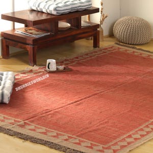 Pode incluir: Um tapete retangular vermelho com uma borda geométrica e franjas. O tapete é feito de um material tecido e tem o texto "DHURRIESWORLDIN". Uma mesa de centro de madeira e uma almofada listrada estão no fundo.