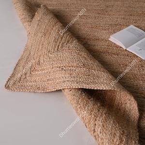 May include: A natural fiber rug made from jute, with a woven texture and a fringed border. The rug is folded in half, revealing the underside.