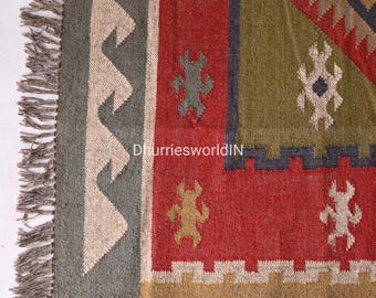 May include: A close-up of a colorful handwoven rug with geometric patterns in shades of red, green, blue, and beige. The rug has fringe on the edges and is made of natural fibers.