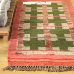 May include: A rectangular rug featuring a geometric design in green, white, and red. It has a pink border and a fringe along the bottom. The text "DHURRIESWORLDIN" is visible at the top of the rug.