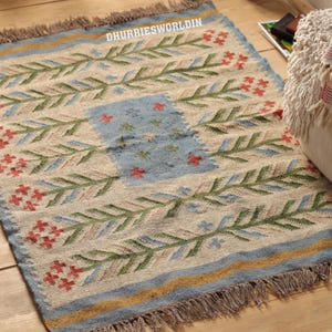 May include: A rectangular woven rug featuring a cream-colored base with a repeating pattern of green leafy branches and small red floral clusters. The center has a light blue rectangular panel, and the edges include blue and yellow borders with fringe.