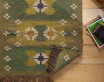 May include: A rectangular woven rug featuring a green and olive base with repeating geometric patterns in white, dark blue, and orange. The rug includes the text DHURRIESWORLDIN and has brown fringe along the bottom edge and one corner.