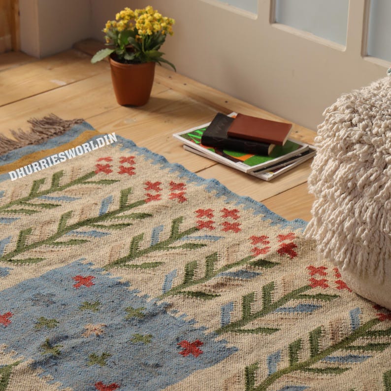 May include: A rectangular woven rug featuring a cream-colored base with repeating green leaf patterns, red floral accents, and a light blue rectangular section. The rug includes the text DHURRIESWORLDIN printed in white on the left side.