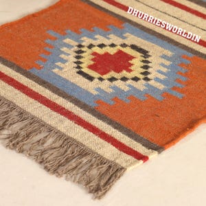 May include: A woven rug featuring a central geometric diamond pattern in red, yellow, and blue on an orange background. The rug includes horizontal stripes in red and grey, finished with a fringed edge. The text DHURRIESWORLDIN is visible.