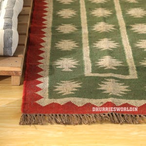May include: A green and red area rug with a geometric pattern. The rug features a central green field with white tree-like designs and a red border with a zig-zag edge. The rug has a fringe along the bottom edge.