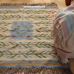 May include: A woven rug featuring a cream base with horizontal rows of green leaf-like patterns, blue accents, and a central blue rectangular panel with small red and green floral motifs. The text DHURRIESWORLDIN is visible. It has brown fringe edges.