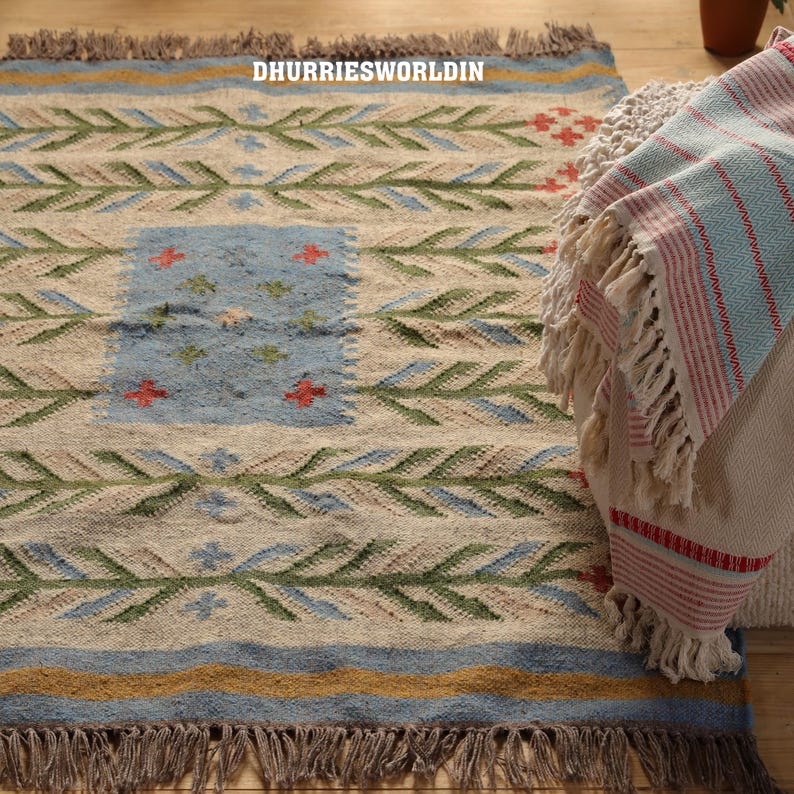 May include: A woven rug featuring a cream base with horizontal rows of green leaf-like patterns, blue accents, and a central blue rectangular panel with small red and green floral motifs. The text DHURRIESWORLDIN is visible. It has brown fringe edges.