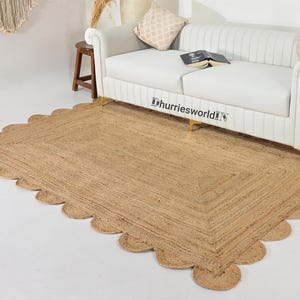 May include: A rectangular jute rug with a scalloped edge is displayed on a white floor. A white sofa with gold legs and two patterned pillows sits on the rug. A small wooden side table and a woven wall hanging are also visible.