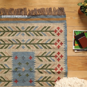 May include: A rectangular woven rug featuring a natural beige base with a repeating pattern of green, blue, and tan leaf-like motifs and red cross-shaped accents. The rug has a blue border, a mustard yellow stripe, and brown fringe at the top edge.