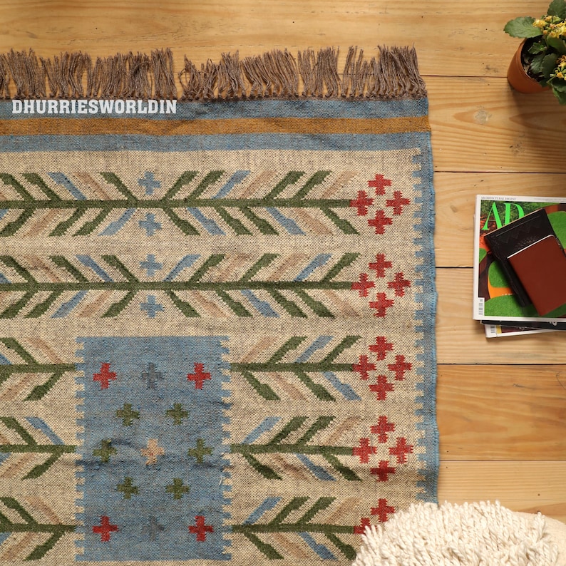 May include: A rectangular woven rug featuring a natural beige base with a repeating pattern of green, blue, and tan leaf-like motifs and red cross-shaped accents. The rug has a blue border, a mustard yellow stripe, and brown fringe at the top edge.
