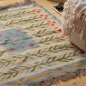 May include: A woven area rug featuring a cream-colored base with green leaf patterns, red floral accents, and a central light blue rectangular section. The rug includes the text DHURRIESWORLDIN, a blue and yellow border, and fringed edges on the ends.