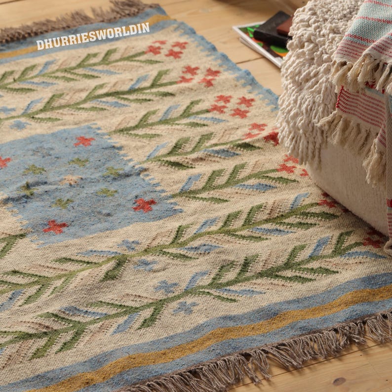 May include: A woven area rug featuring a cream-colored base with green leaf patterns, red floral accents, and a central light blue rectangular section. The rug includes the text DHURRIESWORLDIN, a blue and yellow border, and fringed edges on the ends.