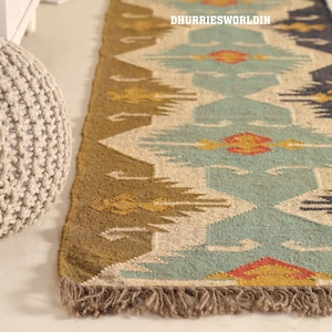 May include: A rectangular rug with a geometric pattern in shades of teal, brown, yellow, and red. The rug has a fringed edge and the text "DHURRIESWORLDIN" is visible. The rug is placed on a light-colored surface.