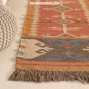 May include: A rectangular woven rug featuring a terracotta red centre with geometric diamond patterns in cream and teal. The border displays a dark brown section with blue and red motifs, finished with a fringed edge. The text DHURRIESWORLDIN is visible.