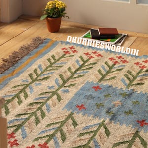 May include: A rectangular woven rug featuring a beige base with repeating green and blue floral patterns and red accents. The rug has a fringed edge and the text DHURRIESWORLDIN is printed in white across the upper portion of the fabric surface.