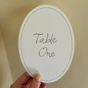 May include: An oval, off-white table number card with the words "Table One" in elegant black script. The card has a thin, black border and a textured surface. Suitable for weddings or events.