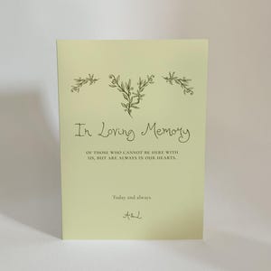 May include: A light green card with the text "In Loving Memory" and a message of remembrance. Two silver rings are in the foreground. The card has floral illustrations and the text "Today and always."