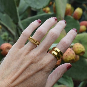 Könnte beinhalten: Zwei goldene Ringe an einer Hand. Ein Ring ist ein dicker, klobiger Kettenglied-Stil. Der andere Ring ist ein einfaches, breites Band.