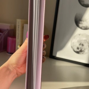 May include: A close-up of the spine of a book with a light purple cover and gold accents. The book is closed and the pages are visible.