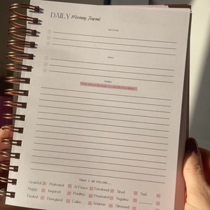 May include: A daily morning journal with a pink and white cover. The journal has a spiral binding and lined pages. The text on the cover reads "DAILY Morning Journal". The journal has sections for gratitude, goals, and a journal entry. The journal also has a section for tracking your mood with checkboxes for feelings like grateful, happy, excited, motivated, inspired, energized, at peace, positive, calm, emotional, tired, frustrated, anxious, negative, and stressed.