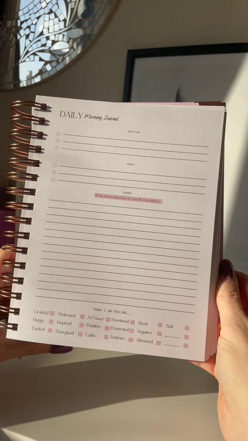 May include: A daily morning journal with a pink and white cover. The journal has a spiral binding and lined pages. The text on the cover reads "DAILY Morning Journal". The journal has sections for gratitude, goals, and a journal entry. The journal also has a section for tracking your mood with checkboxes for feelings like grateful, happy, excited, motivated, inspired, energized, at peace, positive, calm, emotional, tired, frustrated, anxious, negative, and stressed.