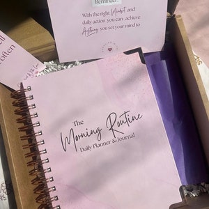 May include: A pink and white marble-patterned journal with a copper spiral binding. The cover reads "The Morning Routine Daily Planner & Journal".