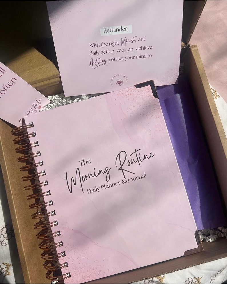 May include: A pink and white marble-patterned journal with a copper spiral binding. The cover reads "The Morning Routine Daily Planner & Journal".