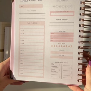 May include: A pink and white daily planner with a rose gold spiral binding. The planner has sections for daily goals, meal planning, water intake, exercise, self-care, and a morning routine tracker. The text on the cover reads "DAILY Morning Planner" and "OWN YOUR MORNING, OWN YOUR LIFE".