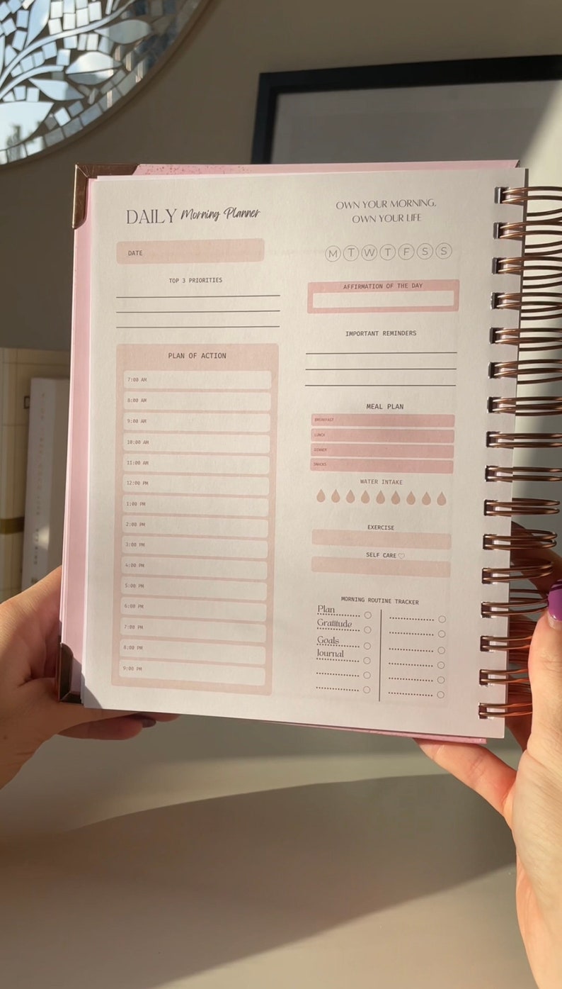 May include: A pink and white daily planner with a rose gold spiral binding. The planner has sections for daily goals, meal planning, water intake, exercise, self-care, and a morning routine tracker. The text on the cover reads "DAILY Morning Planner" and "OWN YOUR MORNING, OWN YOUR LIFE".